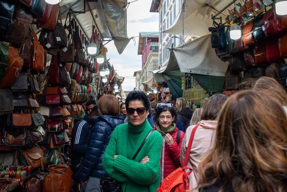 Eye-level view down a bustling, narrow outdoor market alley crowded with people. Stalls on both sides are overflowing with hanging leather bags in various shades of brown. In the foreground, a woman wearing sunglasses and a bright green sweater stands with arms crossed, looking towards the camera amidst the shoppers