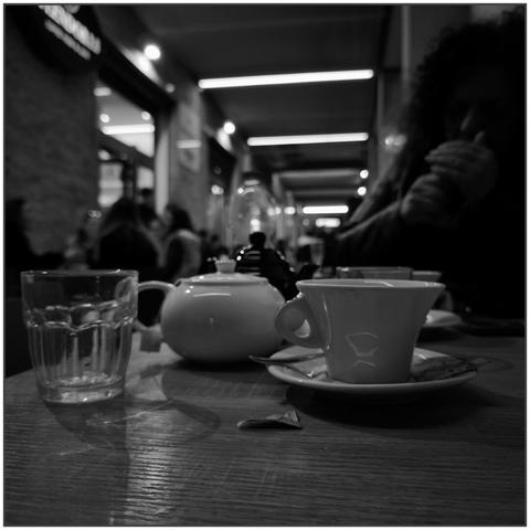 L'immagine è una fotografia in bianco e nero che cattura un momento in un bar o caffetteria. In primo piano, su un tavolo di legno, sono posati una tazza con piattino, una teiera bianca, un bicchiere d'acqua e una bustina di zucchero aperta. La luce riflette delicatamente sulle superfici lisce della ceramica e del vetro.

Sul lato destro dell'immagine, leggermente sfocata, una persona con capelli ricci e voluminosi è intenta a mangiare un panino o un dolce, con le mani che avvolgono l'alimento. Sullo sfondo, sfocato e immerso in un gioco di luci e ombre, si intravede un corridoio illuminato con soffitti a pannelli luminosi, e la presenza di altre persone sedute o in movimento, creando un'atmosfera vivace ma intima.

L'insieme della composizione trasmette una sensazione di calore quotidiano, un momento di pausa e contemplazione in mezzo alla frenesia della vita. Il bianco e nero enfatizza le texture, i contrasti e le luci, aggiungendo un tocco nostalgico e cinematografico alla scena.