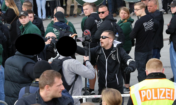 Während Polizeibeamt*innen hinter einem Absperrgitter stehen, greift jenseits des Gitters der offensichtlich dabei auch rufende oder schreiende Philipp Rang mit dem rechten Arm zwei Personen an, die ein Kamerastativ halten. Rang trägt Sonnenbrille, Jacke mit Parteilogo, eine Ordnerbinde und Handschuhe. Ein weiterer Neonazi mit Glatze und Sonnenbrille greift ebenfalls an. Im Hintergrund schauen die TeilnehmerInnen des Aufmarschs in schwarzer oder grüner Parteikleidung zu.
