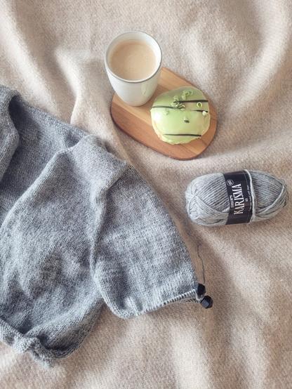 DEU: Ein grauer Strickcardigan in Arbeit, daneben eine schwarzer Kaffee in einem Becher und ein Pistazien-Doughnut mit hellgrüner Glasur

ENG: A grey hand knit cardigan in progress. Next to the knitting project a black coffee and pistachio doughnut