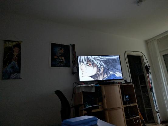 Das Bild zeigt ein dunkles Zimmer mit einem Fernseher, der eine Szene aus einem Anime anzeigt. Der Fernseher steht auf einem Holzregal, das verschiedene Gegenstände wie Kabel und ein weißes Tuch enthält. Links an der Wand hängen zwei Poster: eines zeigt eine Figur mit dem Titel "Trafalgar Law" und das andere ist dunkler mit einem abstrakten Muster. Vor dem Fernseher steht ein schwarzer Bürostuhl, und auf einem Tisch im Vordergrund liegt ein blaues Objekt. Die Beleuchtung im Raum ist gedämpft, was die Szene insgesamt dunkel und intim wirken lässt.

Die Figur auf dem Fernseher hat rote Augen und wird von der Seite gezeigt. Er hat als Haarfarbe eine Mischung aus Weiß und Schwarz.