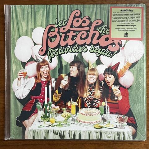 Let The Festivities Begin~ front cover. Featuring four women, the gand members of Los Bitchos, seated in front of white balloons and behind a table set with a cake and party drinks, they are raising glasses and wearing party hats, illustrating the title of "let the festivities begin!"