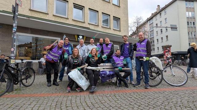 Gruppenfoto von Voltas auf dem Merianplatz in Frankfurt am Main. Die meisten haben Kleidungsstücke im typischen violett und dem Volt-Schriftzug an. Zwei Frauen tragen schwarze Hoodies mit dem Volt-Europa-Logo, bestehend aus dem Volt-Schriftzug in einem Kranz bunter Sterne. Zwei Männer im HIntergrund tragen hellgraue Hoodies.
Die Gruppe bildet einen bunten und fröhlichen Kontrast zum grau verhangenen Himmel und der trist wirkenden Umgebung.