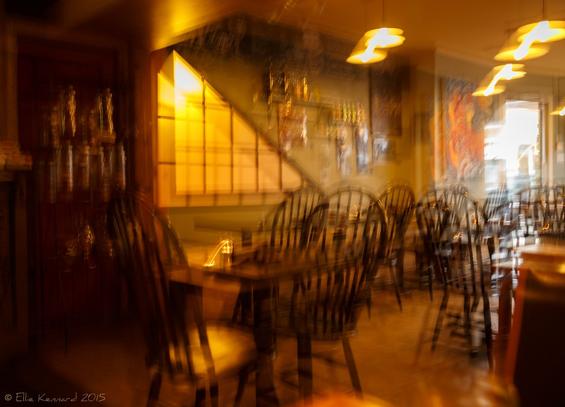A blurred cafe scene with curved back wooden chairs and tables. The lighting is a warm glow and everything is almost double vision. Three hanging lights are turned into 6 lights. There is a triangle of lights with lines through it (doubled) on the wall facing us. The cafe is empty, waiting.