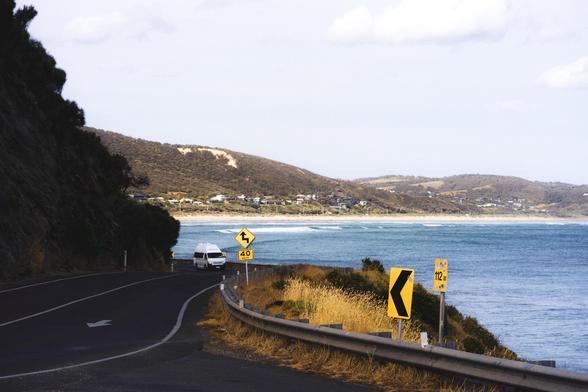 The image shows a coastal road with a vehicle driving along it. The road has a curve and is lined with guardrails. There are several road signs indicating a sharp turn and a speed limit of 40. To the right, there is an expanse of water, likely the ocean, and in the background, there are hills with scattered houses. The setting appears to be a scenic coastal area.