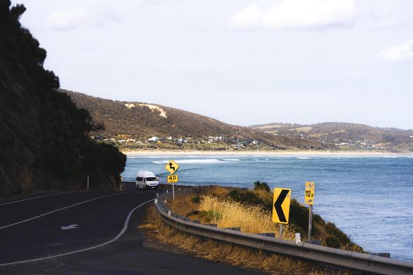 The image shows a coastal road with a vehicle driving along it. The road has a curve and is lined with guardrails. There are several road signs indicating a sharp turn and a speed limit of 40. To the right, there is an expanse of water, likely the ocean, and in the background, there are hills with scattered houses. The setting appears to be a scenic coastal area.
