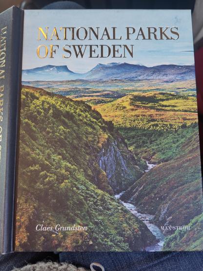 Front cover of a book embossed with gold title National Parks of Sweden showing a photograph of a view over Abisko national park with low canyon running through forest and on the horizon a wide u-shaped Valley cut into high mountain plateau