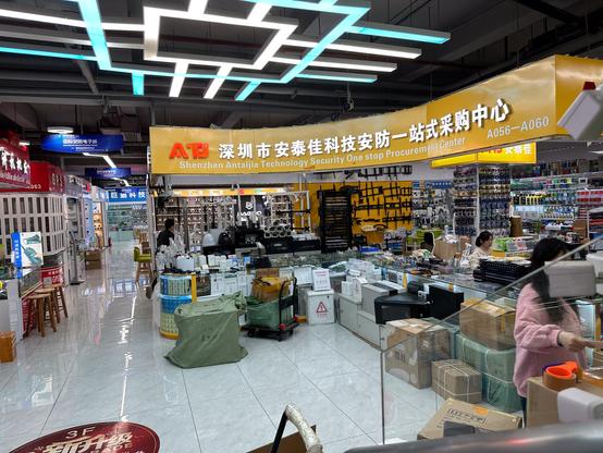 Blick in die Mall. An der Ecke ein Lden mit Überwachungskameras, Halterungen für diese und abschließbare Kästen in der zusätzliche Elektronik der anlagen gebaut werden kann. Vor dem Geschäft viele Kartons und Kisten in denen Angelieferte oder zu versendende Produkte stecken.
