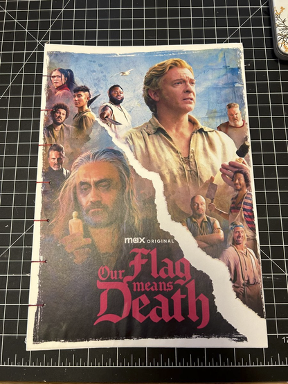 A handbound book of Our Flag Means Death sheet music. The cover is an OFMD Season 2 poster created by xenantis, showing Ed, some of his S2 crew and Zheng on the left, and Stede & his own crew on the right. They are separated by a white strip that runs diagonally across the poster like a rip in the paper.