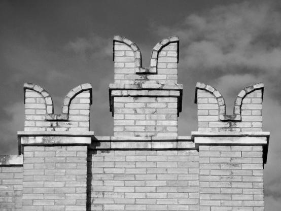 Black and white photo of the crenellated parapet of a light-colored brick building. The parapet features three upright sections, each terminating in a swallowtail.