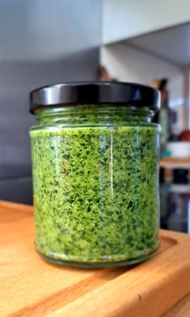 On a wooden board, a clear glass jar with a black lid. In the jar is a bright green substance, flecked with darker green. It is a pesto of wild garlic leaves.