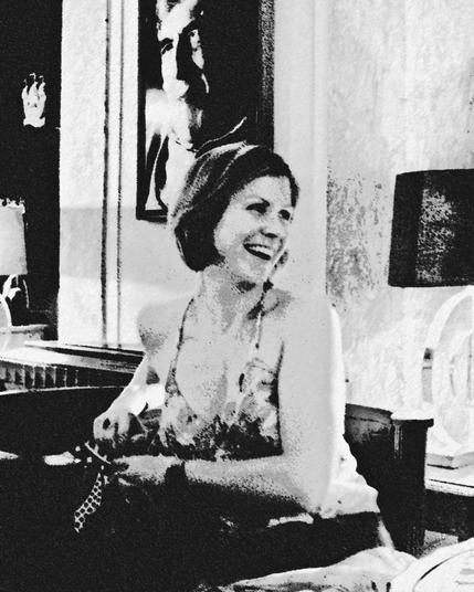 A black and white image of a woman sitting and laughing, wearing a patterned sleeveless dress. In the background, there is a framed photograph of a man. The setting is indoors at hotel bar/lounge, with visible table lamps and other furnishings.