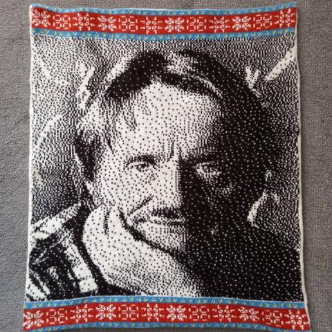 Knitted panel showing a black and white, pixelated, head and shoulders portrait of a white man looking straight at the viewer. He is resting his chin on one hand and is wearing a checked shirt.

Above and below the portrait are bands of colourful motifs. Narrow blue stripes with yellow gliders on either side of red stripes with white pulsars.