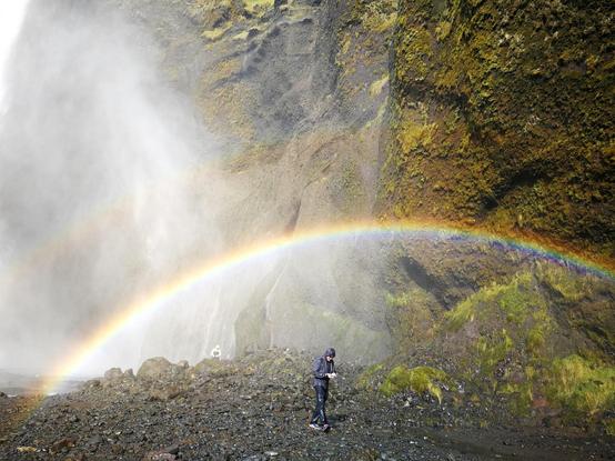 A picture of a misty mountain side with a waterfall on the left and a rainbow across the center. There's a person standing in the middle below the rainbow