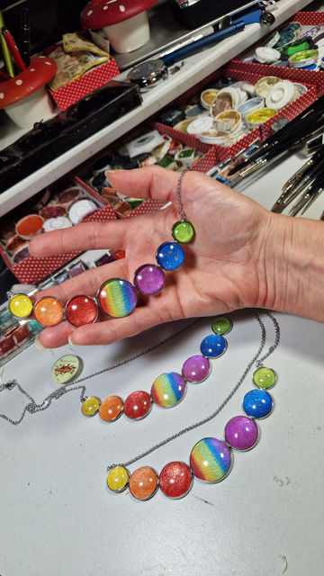 Halsschmuck, Edelstahl, groß, mit 7runden  abgestuft aneinandergereihten handgemalten unter Glas gefassten Motivfarbsteinen in  Regenbogenfarben, an meinem Arbeitstisch fotografiert, einer in der Hand ,2 weitere liegen daneben