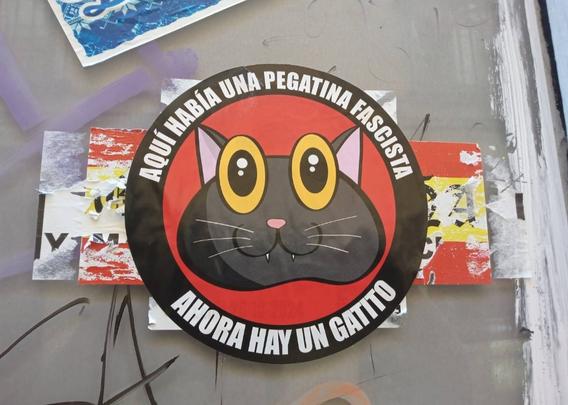 Pegatina de la graciosa cabeza de un gato negro de ojos saltones de color amarillo.
Forma circular con bordes negros y fondo rojo.
En el borde pone "Aquí antes había una pegatina fascista. Ahora hay un gatito"la pegatina está tapando unas pegatinas fascistas españolas.