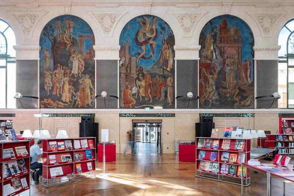 Interior of a Neoclassical Library with three large blind arches surrounding coloured frescoes. The furniture is contemporary in style.