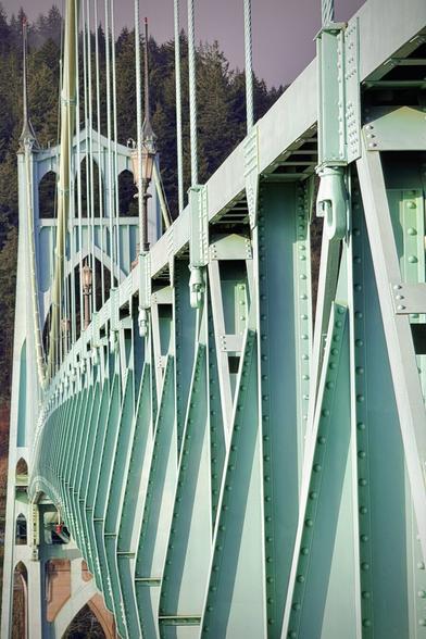 Consecutive sea-foam-green suspension bridge cables, beams, and trusses are viewed from a left-side diminishing perspective, as they lead off into the distance where the second of two towers stands against a cloudy, green-tree-hill backdrop.