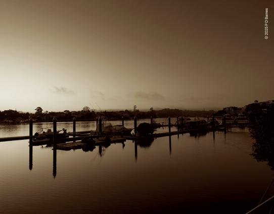 A dawn riverscape, light brightening on the horizon over the far bank on the left, still quite dark further up-river in the distance on the right. The line of the opposite bank is repeated in a group of boat moorings on the near bank. The sky is clear, the river is calm, the day begins.