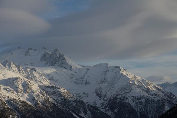 Die Mischabelgruppe im Abendlicht von der Moosalp her gesehen. verschneite felsige Gebirgsrücken umschliessen einen Gletscher. Darüber geschwungene und etwas verwehte Föhn-Fische. Das sind geschwungene Wolken mit schönen abgegrenzten Formen.