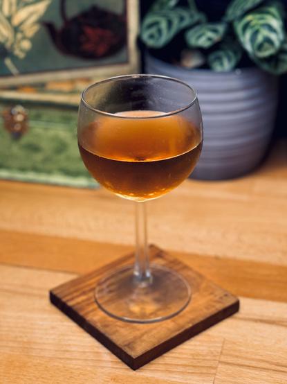 golden cocktail in a chilled coupe on an olive wood coaster with a tea box and plant in the background