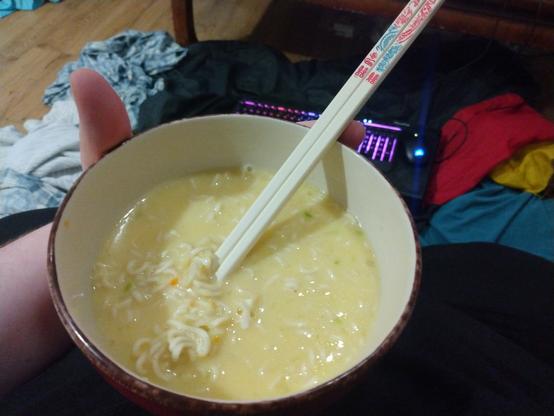 A bowl full of instant ramen with a pair of plastic chopsticks poking out of it. It doesn't look like much, but it was delicious! My cluttered room is vaguely visible in the background, along with a keyboard and mouse that have RGB lighting.