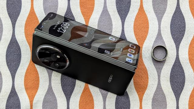 Picture of black Oppo Find N5 book-style folding phone half open (tent mode) next to a silver UltraHuman Ring Air smart ring on an orange and grey patterned mid-century modern placemat