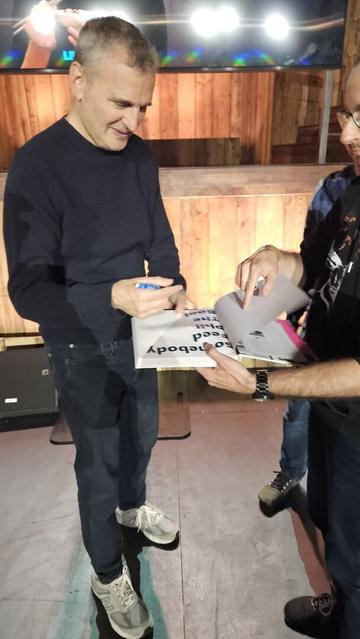 Photo of Phil Rosenthal signing my copy of his book. He's very tall and wears black comfy looking clothes. I'm at the edge of the photo holding the book. We're on a wooden stage. Bright spotlights are reflected on a screen in the background.