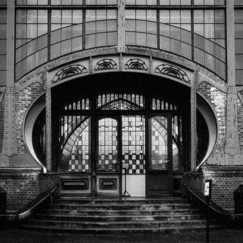 Black-and-white photograph of an Art Nouveau-style entrance of an industrial building with intricate ironwork, curved lines, and stained glass windows arranged in a geometric pattern. The symmetrical composition features an arched doorway with ornate eye-like motifs above it, flanked by circular brick and metal structures.
