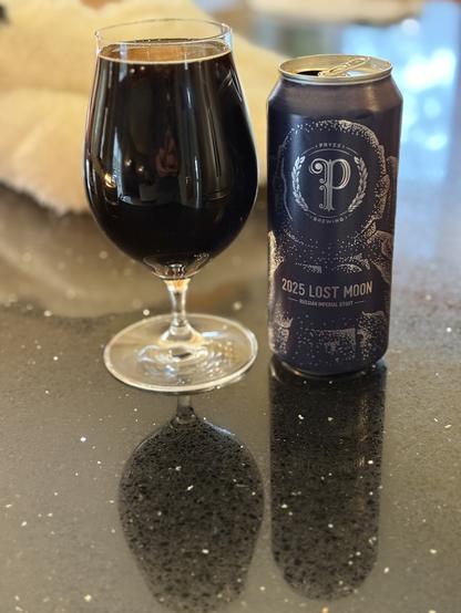 A tall snifter glass of dark beer. To the right is a dark blue can with silver lettering, a large scripted “P” for the Pryes logo, and the abstract outline of an astronaut in a space suit.