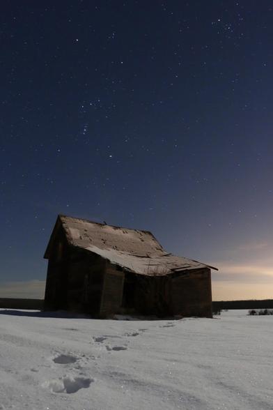 A delapidated old farm house stands in a snowy field, footprints leading towards the house from the camera. In the night sky above, stars are visible, including the constellation of Orion.