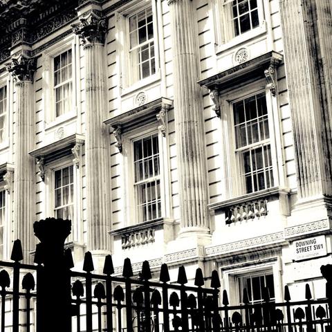 The image shows a close-up of a classical-style building facade with tall columns and large windows. The structure features ornate details, including decorative carvings above the windows and along the edges of the columns. In the foreground, there is a wrought-iron fence with pointed tips. A sign indicating "Downing Street SW1" is visible.