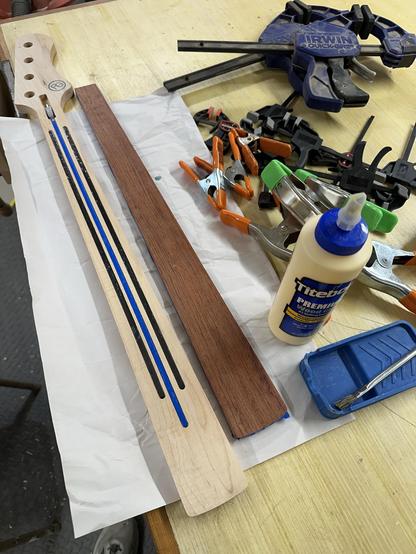 An image of a workspace featuring wood pieces, including a guitar neck with inlays and a fretboard, a bottle of wood glue, a paintbrush, and various clamps. The background includes tools and materials arranged on a wooden table.