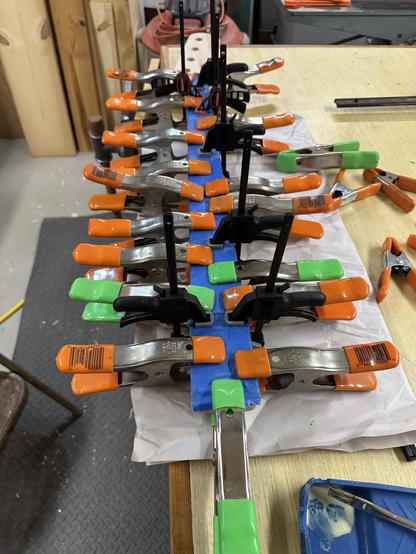 A setup of various clamps with orange and green handles securing a project, positioned over a workbench. Blue painter's tape is visible beneath the clamps, along with a small container and brush for adhesive. The background includes wood pieces and workshop tools.