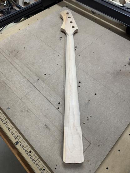 A raw, unfinished bass guitar neck is laid out on a workbench. The neck features a headstock with three drilling holes and has a smooth surface, with visible wood grain. The background includes a work surface with measurement markings and a few tools.