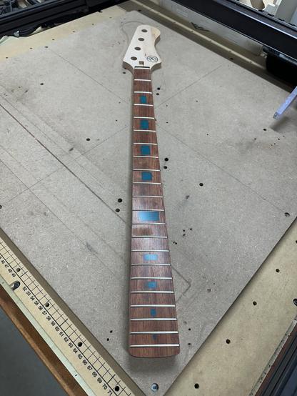 An unfinished bass guitar neck is shown, featuring a wooden fingerboard with inlays and fret markers. The neck has a headstock with pre-drilled tuner holes and is lying on a work surface that includes measurement lines.