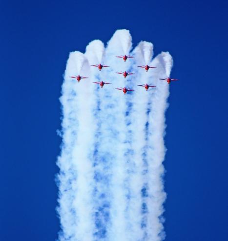 A squadron of nine red aircraft (the Red Arrows) fly in a tight formation, leaving thick white smoke trails behind them against a clear blue sky. The planes are positioned in a diamond shape, with precise alignment and symmetry, demonstrating their aerobatic skills.