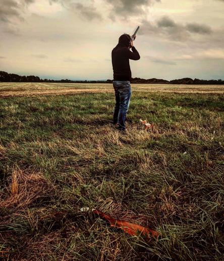A person stands in a field, aiming a shotgun towards the sky. The individual is dressed in a black top and jeans, and the scene is set under a cloudy sky, giving a moody atmosphere. The grassy field stretches out into the distance with trees lining the horizon.