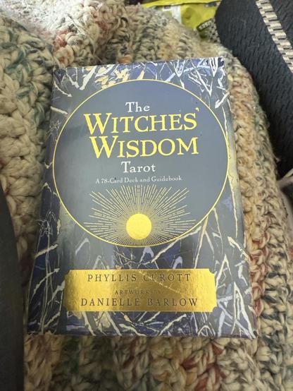 The Witches Wisdom Tarot, a 72 card deck and guidebook, by Phyllis Currott and Illustrated by Danielle Barlow. Box is on a light multicoloured afghan and beside a black couch cushion with a white and grey stripe.