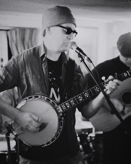 The image shows a musician playing a banjo and singing into a microphone. He is wearing a beanie and sunglasses. The background is slightly blurred, focusing on the performer. Another musician can be partially seen playing an acoustic guitar. The photo is in black and white.