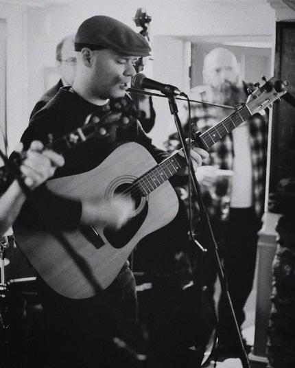 This black and white photo captures a musician playing an acoustic guitar while singing into a microphone. He is wearing a flat cap and a dark sweater. The focus is on the musician in the foreground, while the background is slightly blurred.