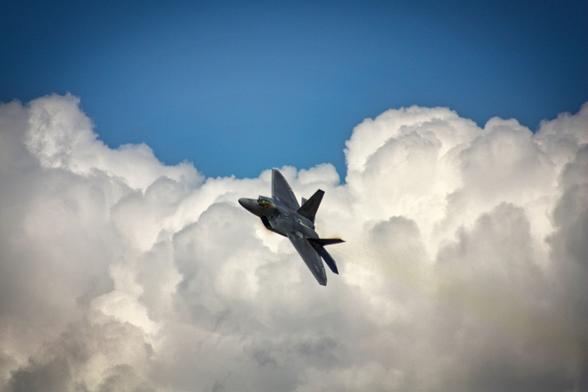 A military jet – the F-22 Raptor – is captured mid-flight against a backdrop of fluffy white clouds and a clear blue sky. The sleek, angular design of the aircraft contrasts sharply with the softness of the clouds, emphasizing its speed and power.