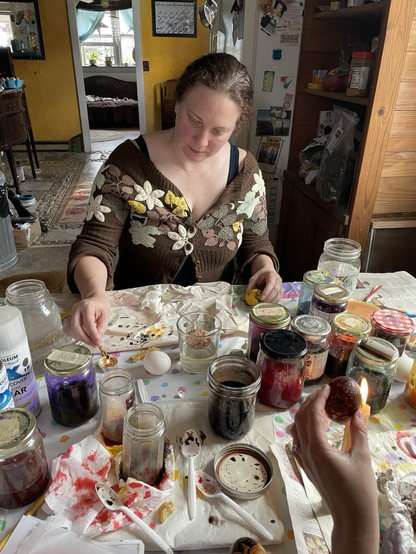 A woman wearing a flower sweater writing pysanky. Eggs, jars of dyes and candles are on a table.