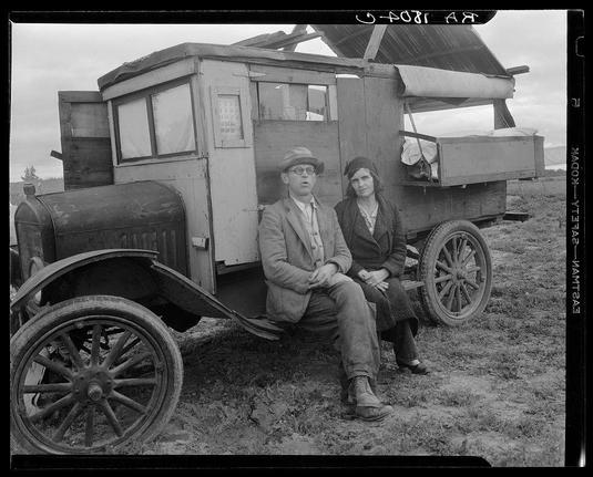 The image depicts a black and white photograph featuring two individuals, likely men and women from the early 1900s era. They are sitting on an old-fashioned truck that appears to be used for agricultural purposes such as transporting goods or farming equipment.

One of them is wearing glasses, a hat, a jacket with buttons, and trousers while holding onto his coat. The other individual wears a dark-colored coat with fur trim around the hood, gloves, and pants. Both individuals appear weary but are maintaining composed demeanors in front of the camera.

The truck has an open structure without doors or windows on its sides, exposing various internal components like shelves filled with items possibly related to farming activities. The vehicle's paint is faded, and there appears to be dirt and rust visible on the metal surfaces.

In the background, a rural landscape can be seen with fields extending into the distance under a cloudy sky. There are trees in the far field which could suggest that this scene takes place near or within an agricultural area.

The photograph includes text at its edges indicating it was taken by Dorothea Lange and references her project on pea pickers in California, dating back to 1938-42. The image captures a moment of rest during the workers' labor-intensive task as they sit amidst their work environment, possibly reflecting upon the challenges faced while earning meager wages.

This photograph pr [...]