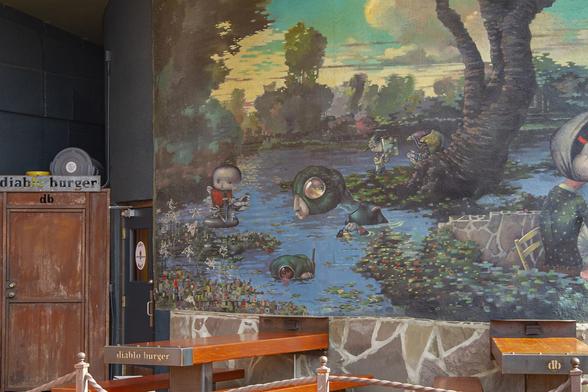 Joe Sorren mural “The Veridic Gardens of Effie Leroux.” 2000 at the patio of Diablo Burger in downtown Flagstaff Arizona.  Early morning quiet view with silence and empty seating accessory to a powerful landscape scene of a pond and lush foliage being enjoyed by the park visitors, surreal setting.