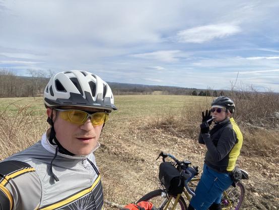 Two cyclists resting and snacking with a hill top farm view around them. It's early spring in New England so there isn't much growing, barely any green showing, and the trees are still bare.