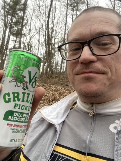 A picture of me after the bike ride, holding a can of Grillos Pickles V8 Dill Pickle Bloody Mary mix which I drank to "replace my electrolytes". There's some trees and brown leaves on the ground behind me because we're in the woods.