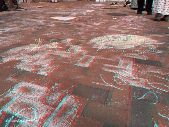 A view of sidewalk chalk art and messages across a large area of brick paving. Red/blue anahlyph view.