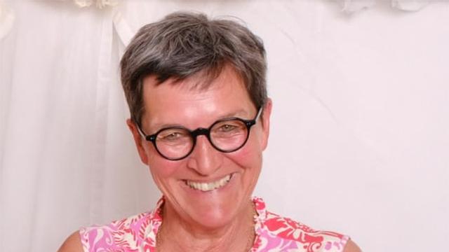 Porträt einer lächelnden Judith Bader-Reissing mittleren Alters mit kurzen, grau melierten Haaren und runden, schwarzen Brille. Sie trägt ein rosafarbenes Oberteil mit Blumenmuster und ist vor einem weißen Hintergrund abgebildet.