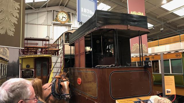 The image shows a historical display in a museum setting, featuring a horse-drawn tram from the 1870s. The tram is brown with a wooden exterior, and a horse is harnessed at the front. The background includes museum signage with the text "1870s Horse Drawn" and an ornate clock on the wall above. Part of the display includes a sign that instructs visitors to "REMAIN SEATED UNDER BRIDGES." There are several people observing the exhibit.